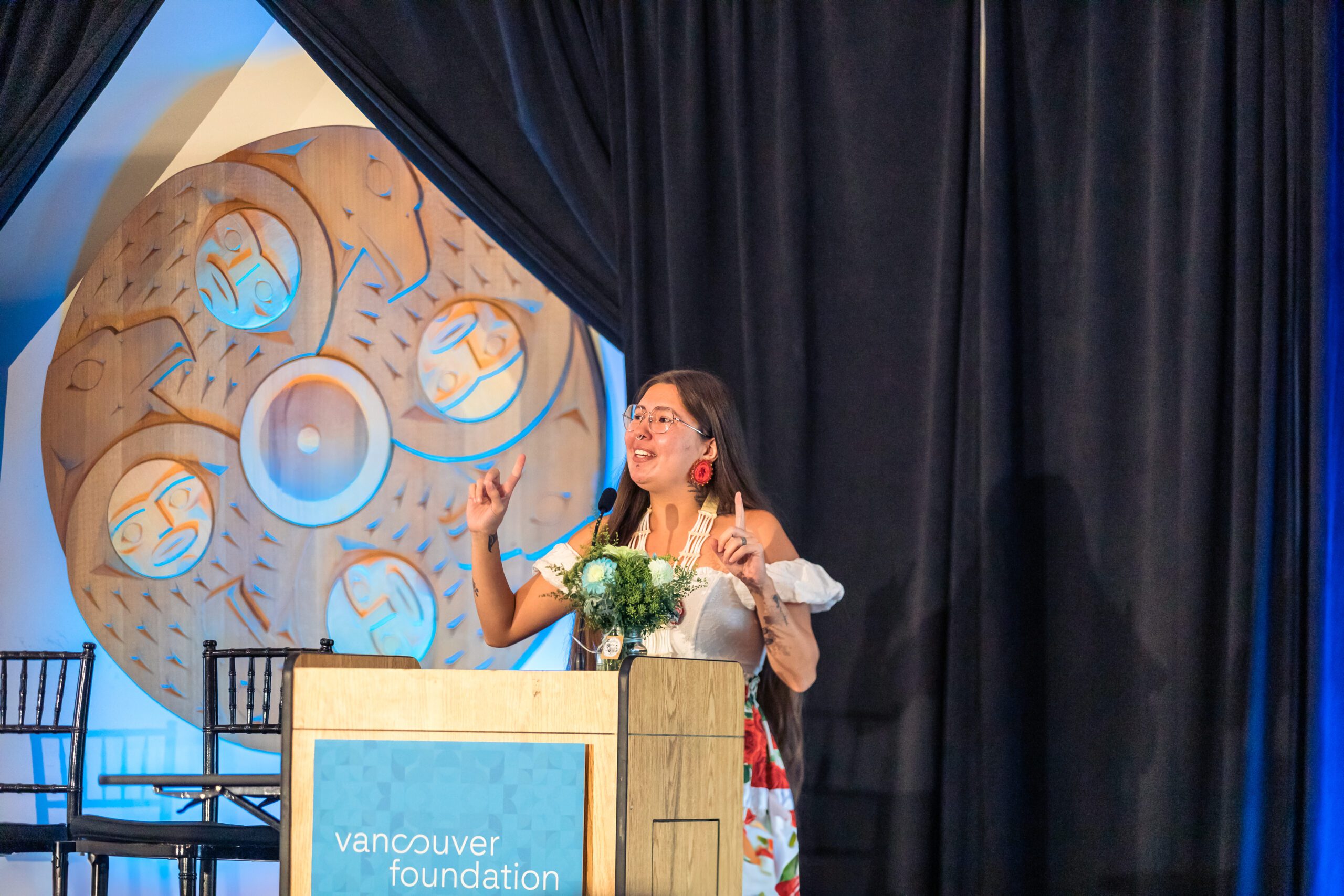 A woman stands behind a podium with the “Vancouver Foundation” logo visible. She has long brown hair and is wearing a floral dress and glasses. Her fingers are pointed up at the ceiling.