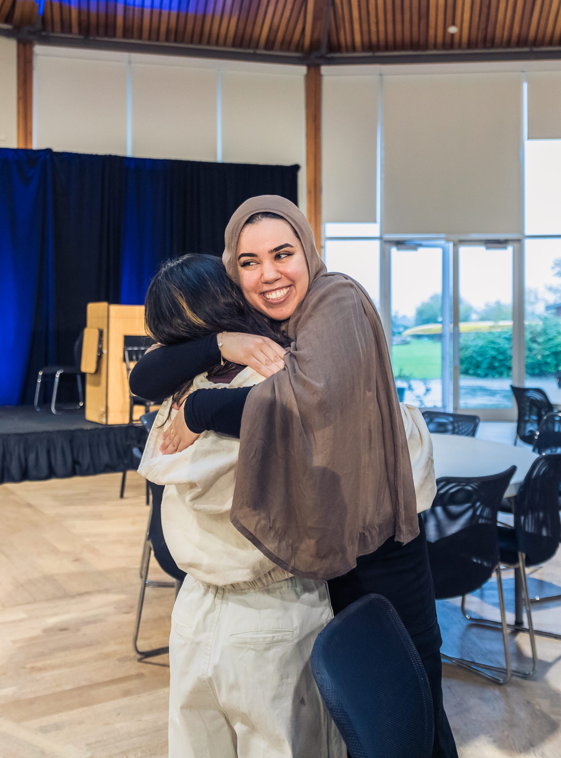 A young woman wearing a hijab hugs another woman with a big smile on her face.