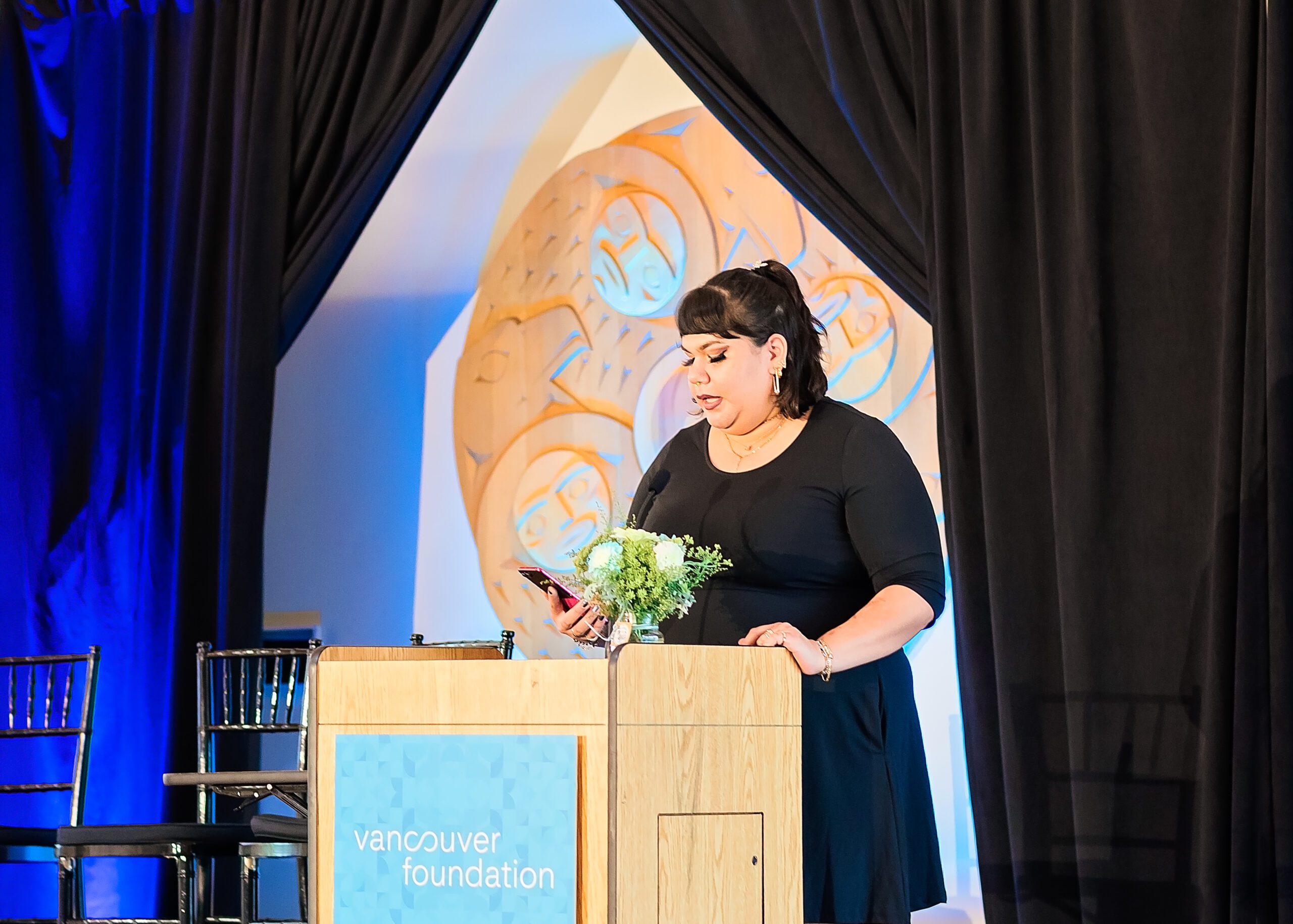 An Indigenous woman stands behind a podium with the “Vancouver Foundation” logo visible. She has shoulder-length black hair and is wearing a black dress. She’s looking down on her phone, which she is carrying in her right hand. On the podium to her right is a vase with a small bouquet of blue and white flowers. Behind her are dark blue curtains.